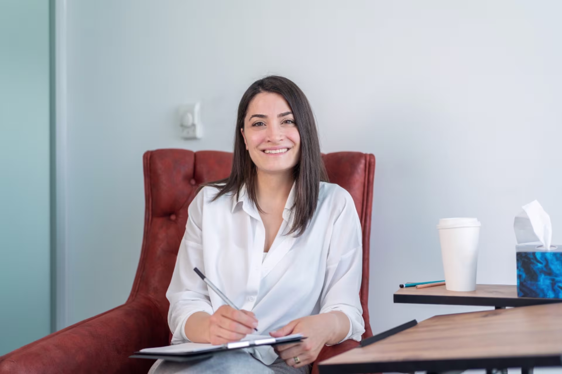 A mental health counselor sits at her desk with notepad in hand smiling at the camera.