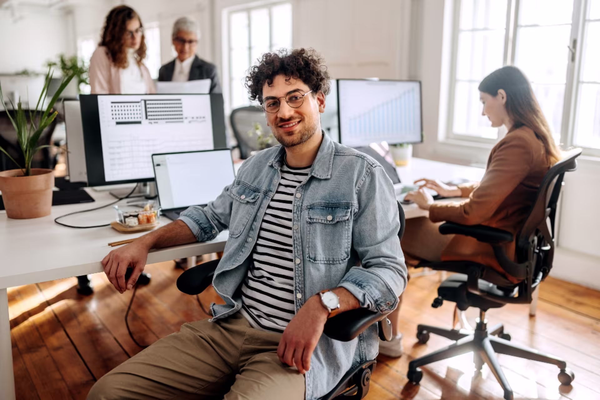 A professional sits at a desk in a collaborative office while colleagues work on computers in the background.