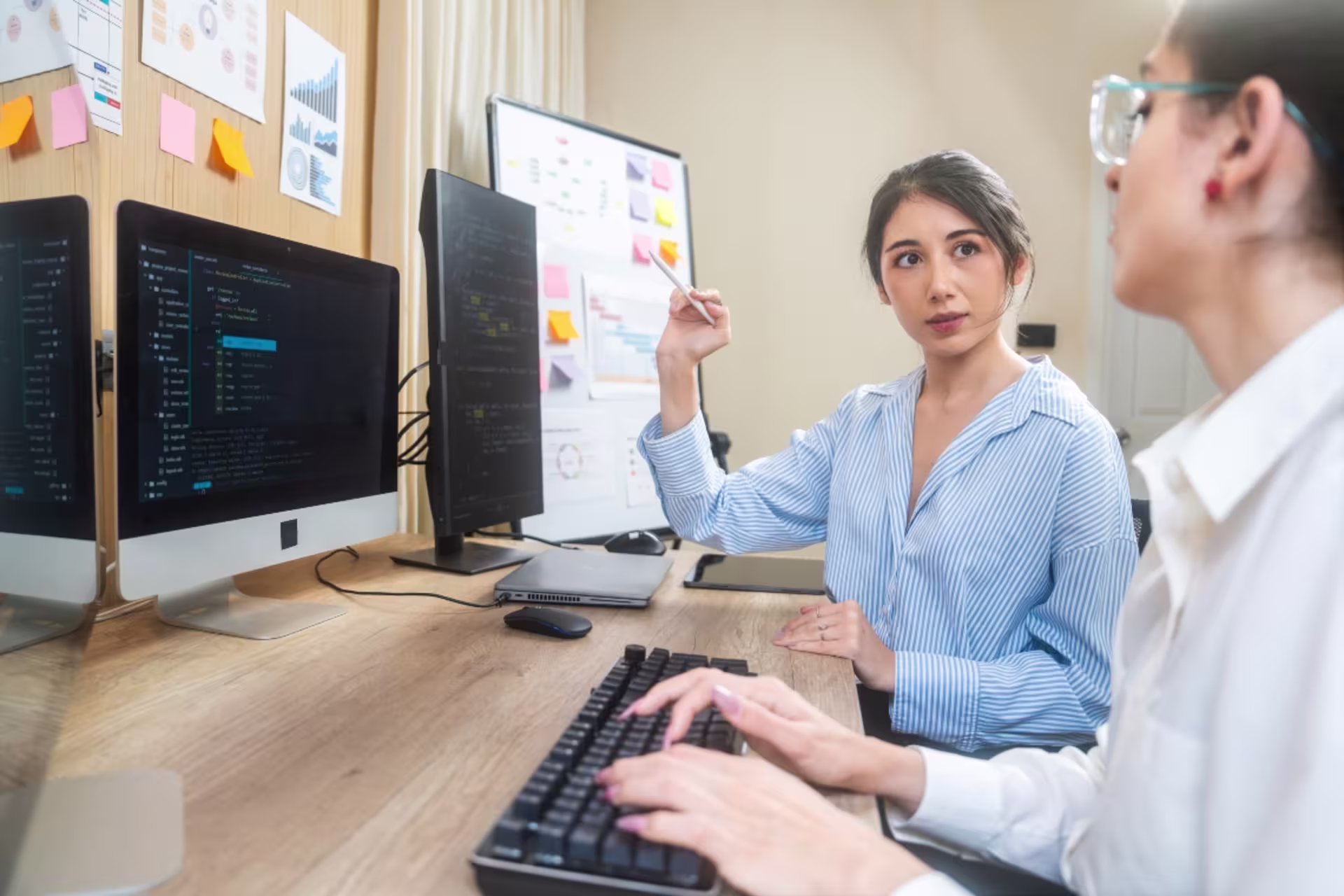 Two women work together at a computer desk, with one pointing toward a monitor displaying code while the other types as they discuss software development tasks.