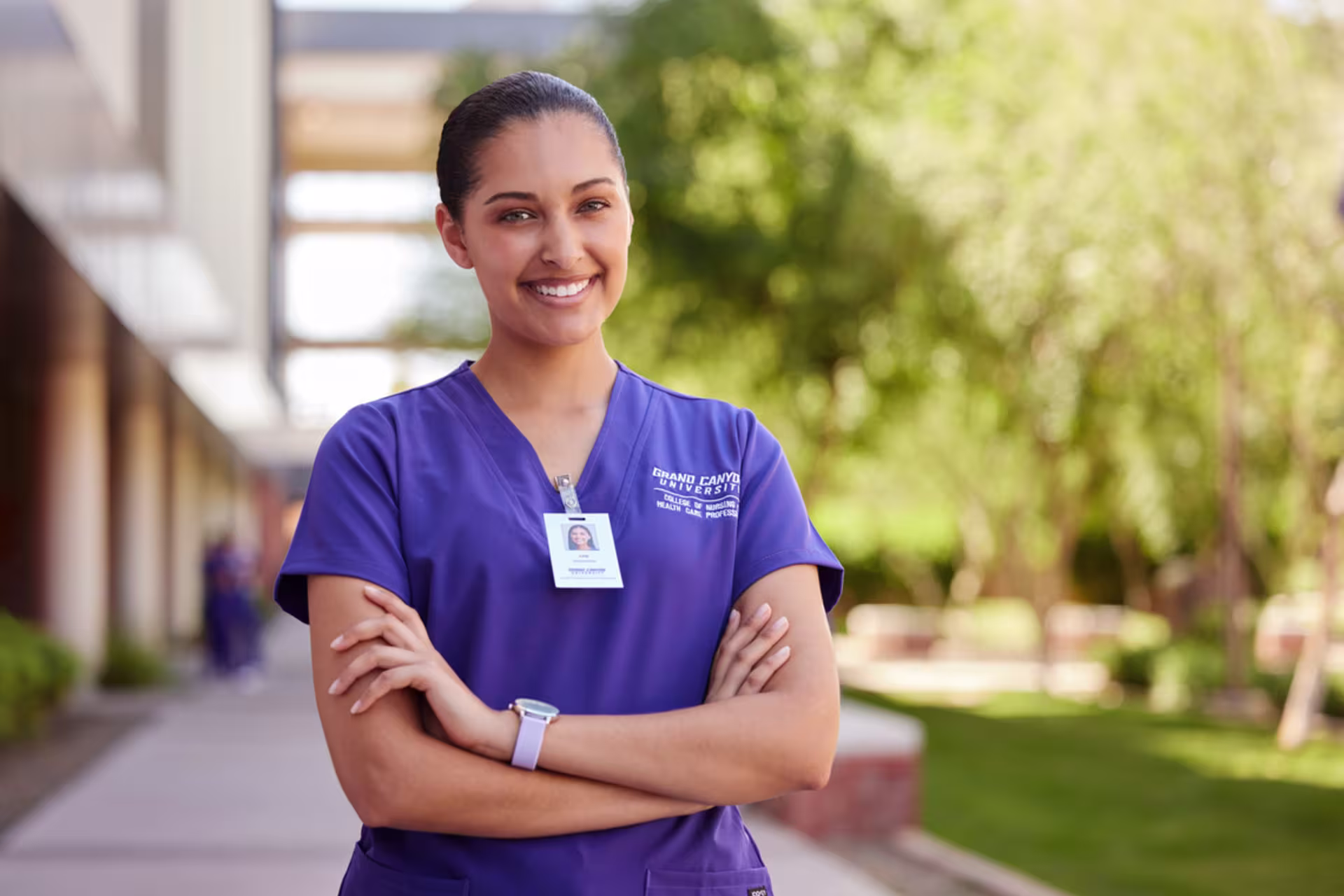 Female nursing student wearing purple scrubs stands with arms crossed outside in a courtyard.