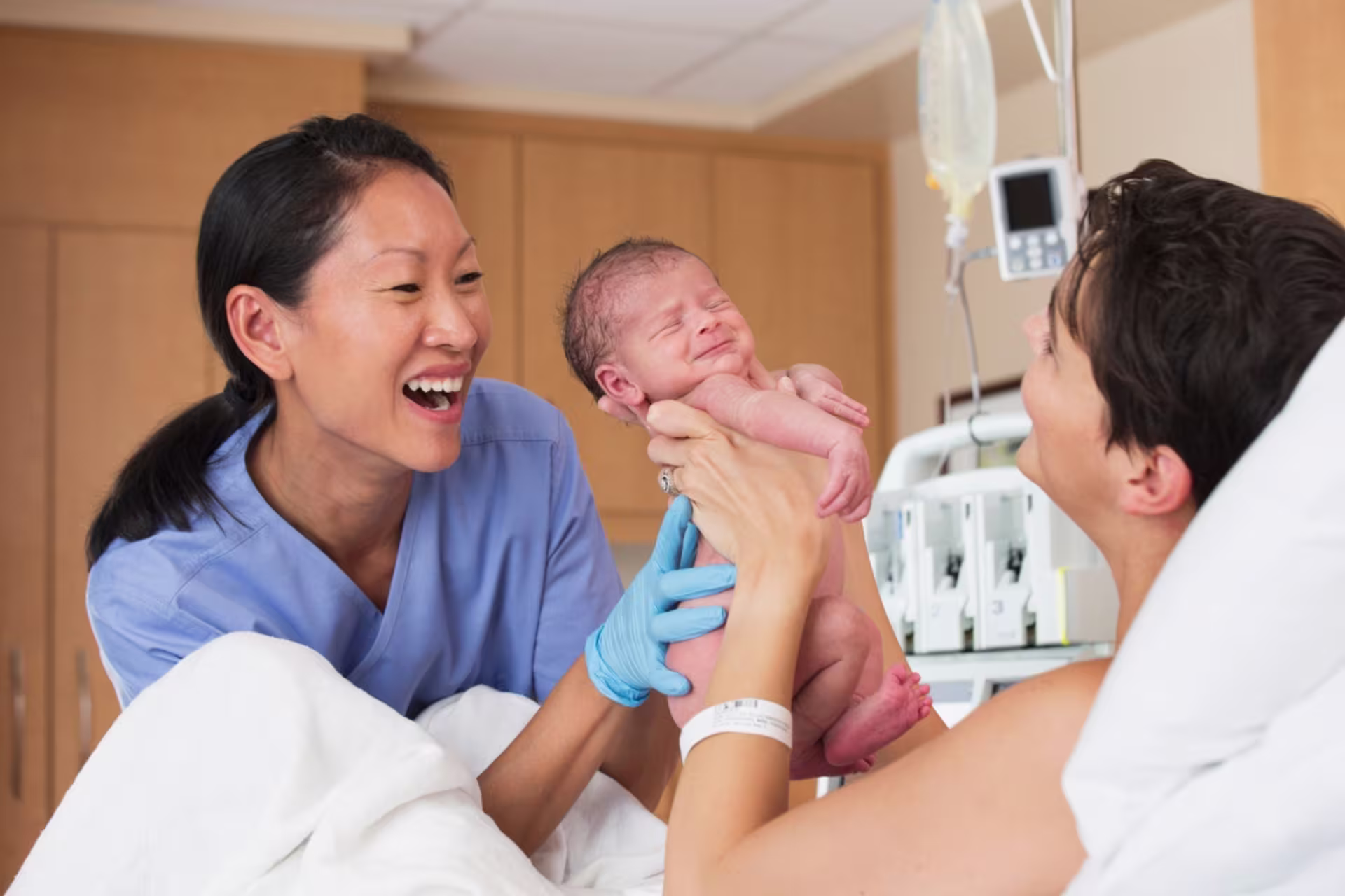 A smiling nurse midwife hands a newborn baby to its mother after delivery in a hospital room.