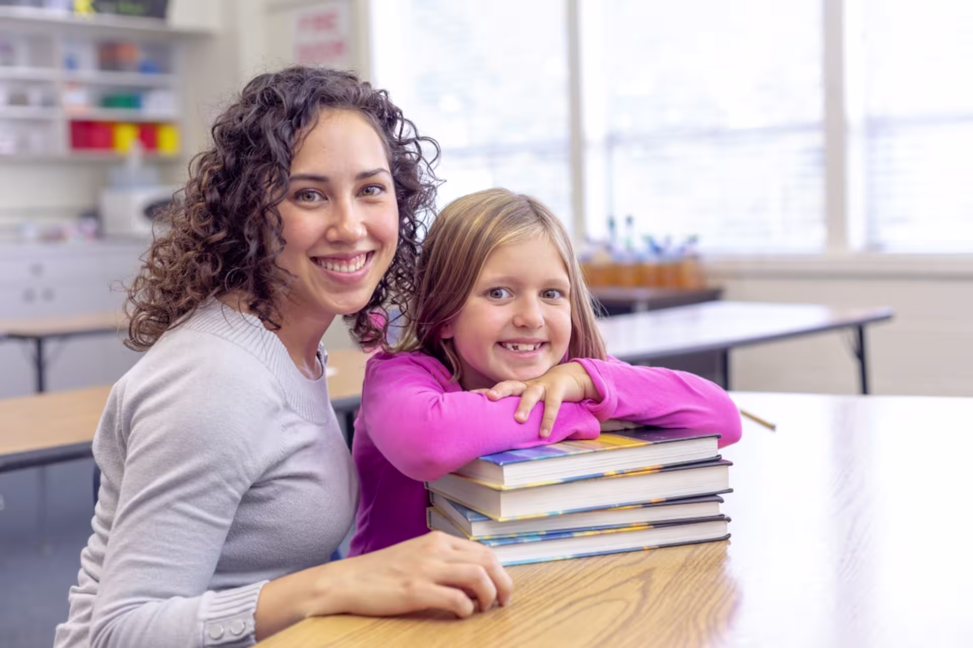 A female reading specialist sits at a table with a young student who is leaning on a stack of books.