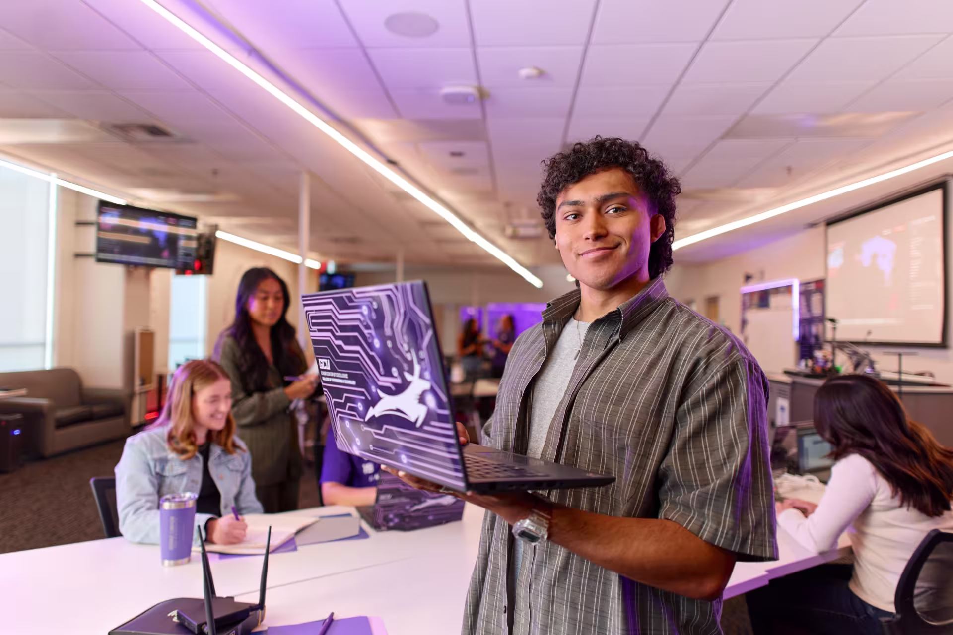 College student in a modern classroom holding a laptop with classmates collaborating in the background.