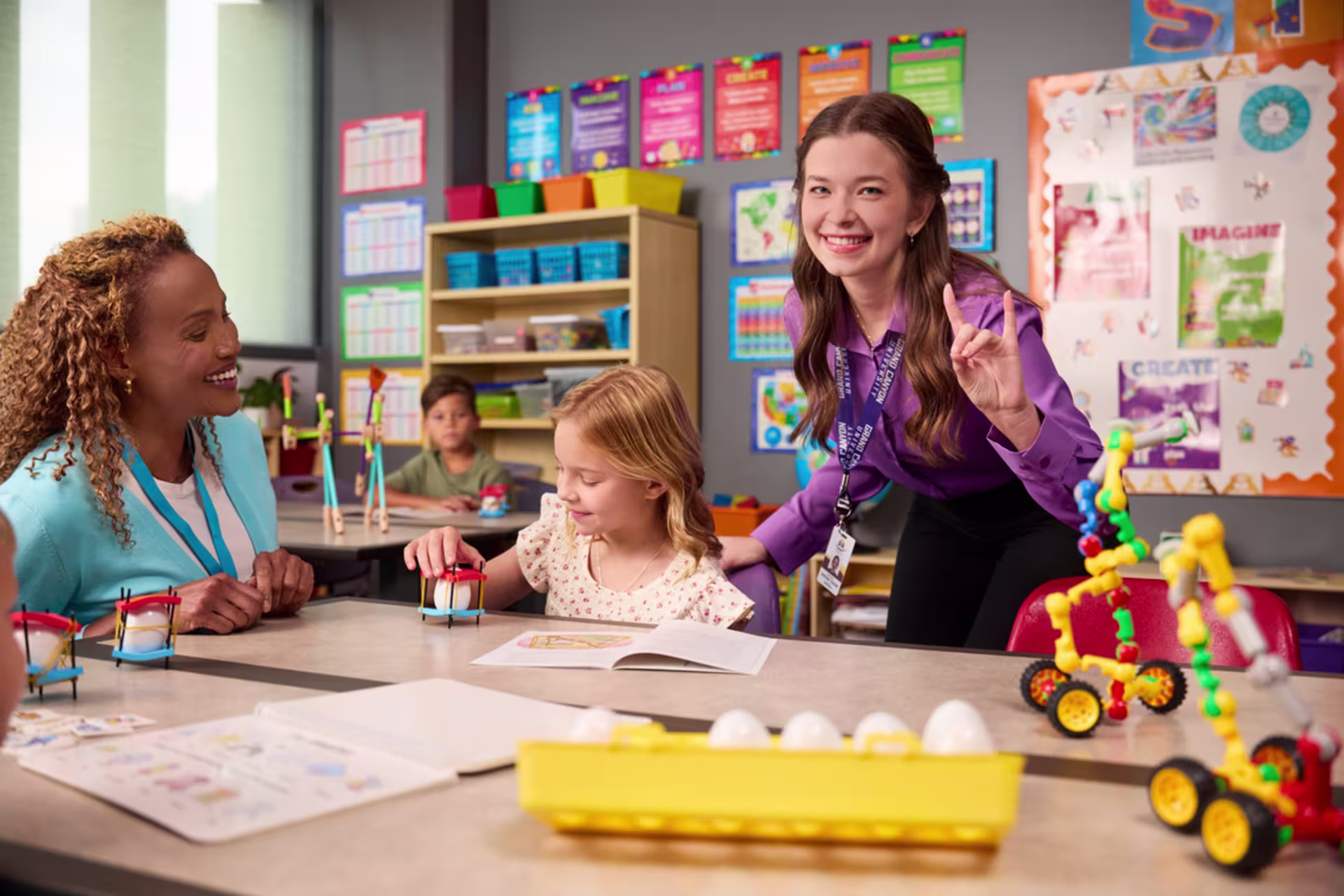 Two female teachers stand in a classroom — one smiles warmly at a student, while the other raises her hand in a gesture of lopes up.