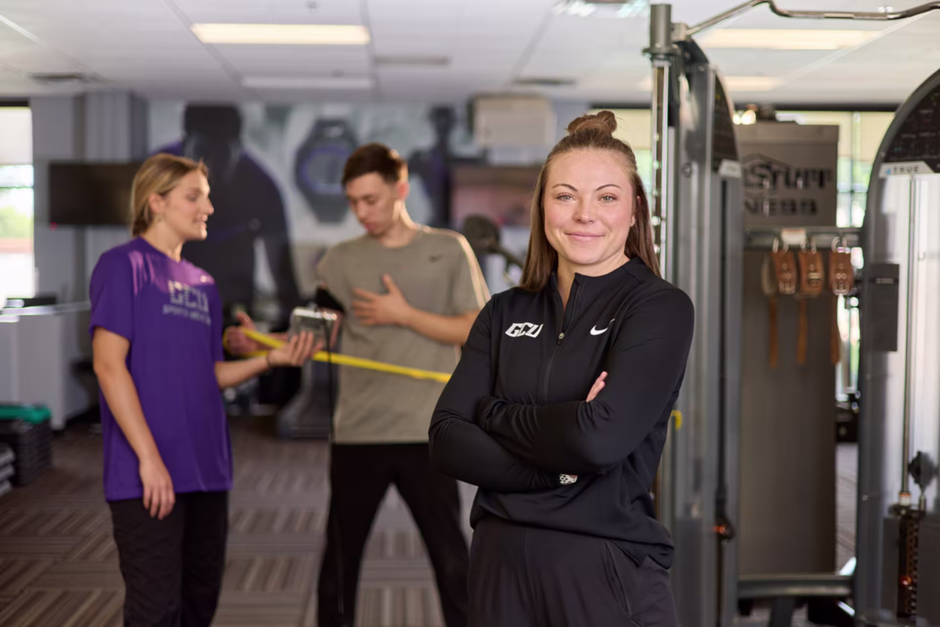 A health scientist stands confidently in a modern gym while clients with resistance band exercise in the background.