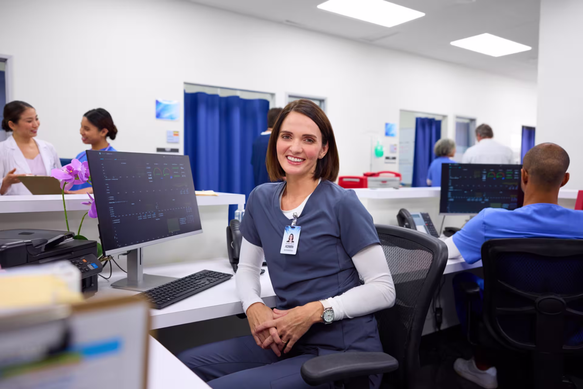 A smiling nurse leader sits in a modern hospital workstation with colleagues working in the background.