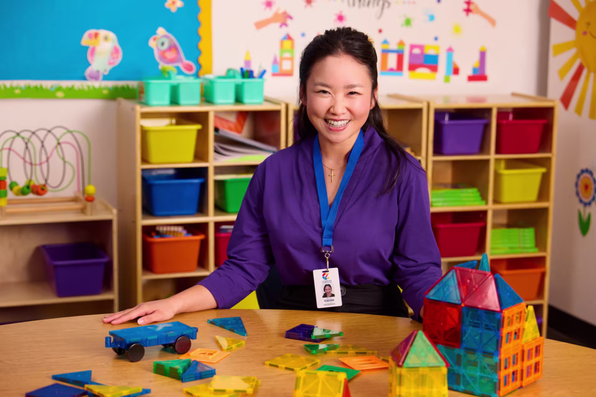 A female preschool teacher sits at a table with colorful toy blocks and smiles at the camera in a classroom setting.
