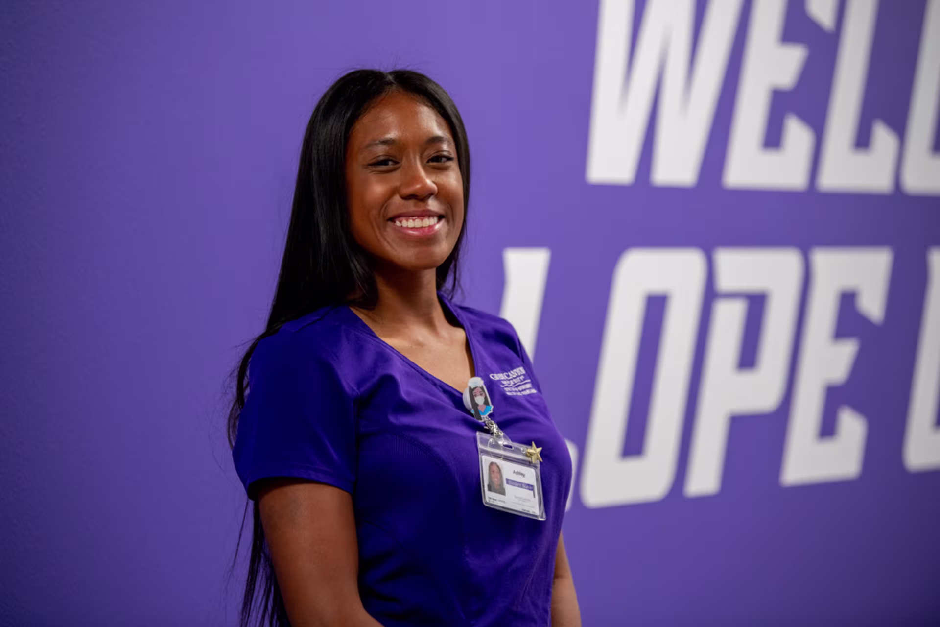 An ABSN in Nevada nursing student wearing purple scrubs stands and smiles in front of a purple wall with white lettering.