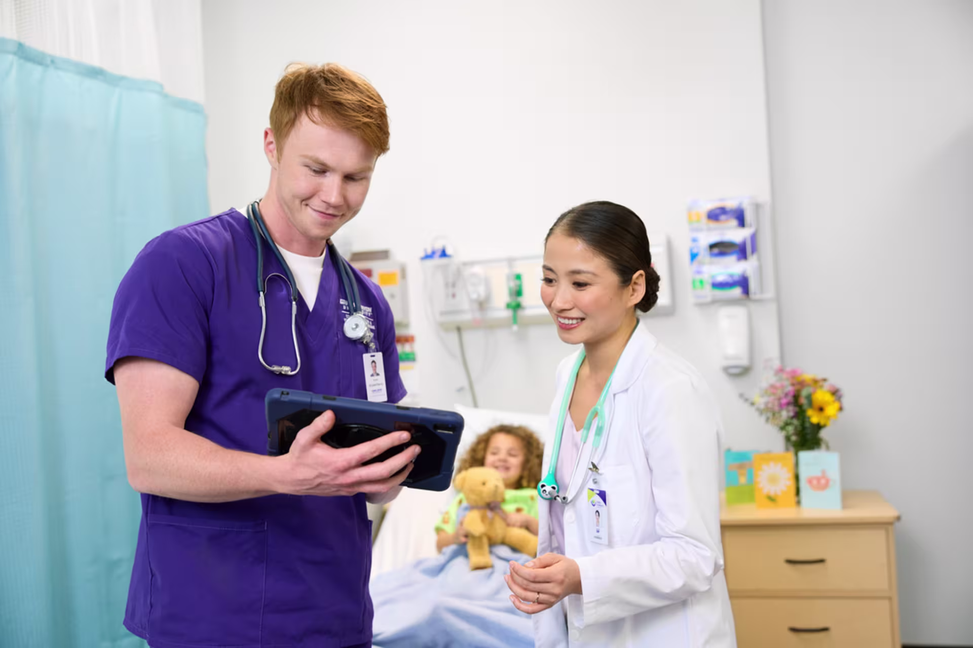 Male nurse in purple scrubs consults with his nursing leader with a female pediatric patient in a hospital bed behind them playing with a stuffed teddy bear.