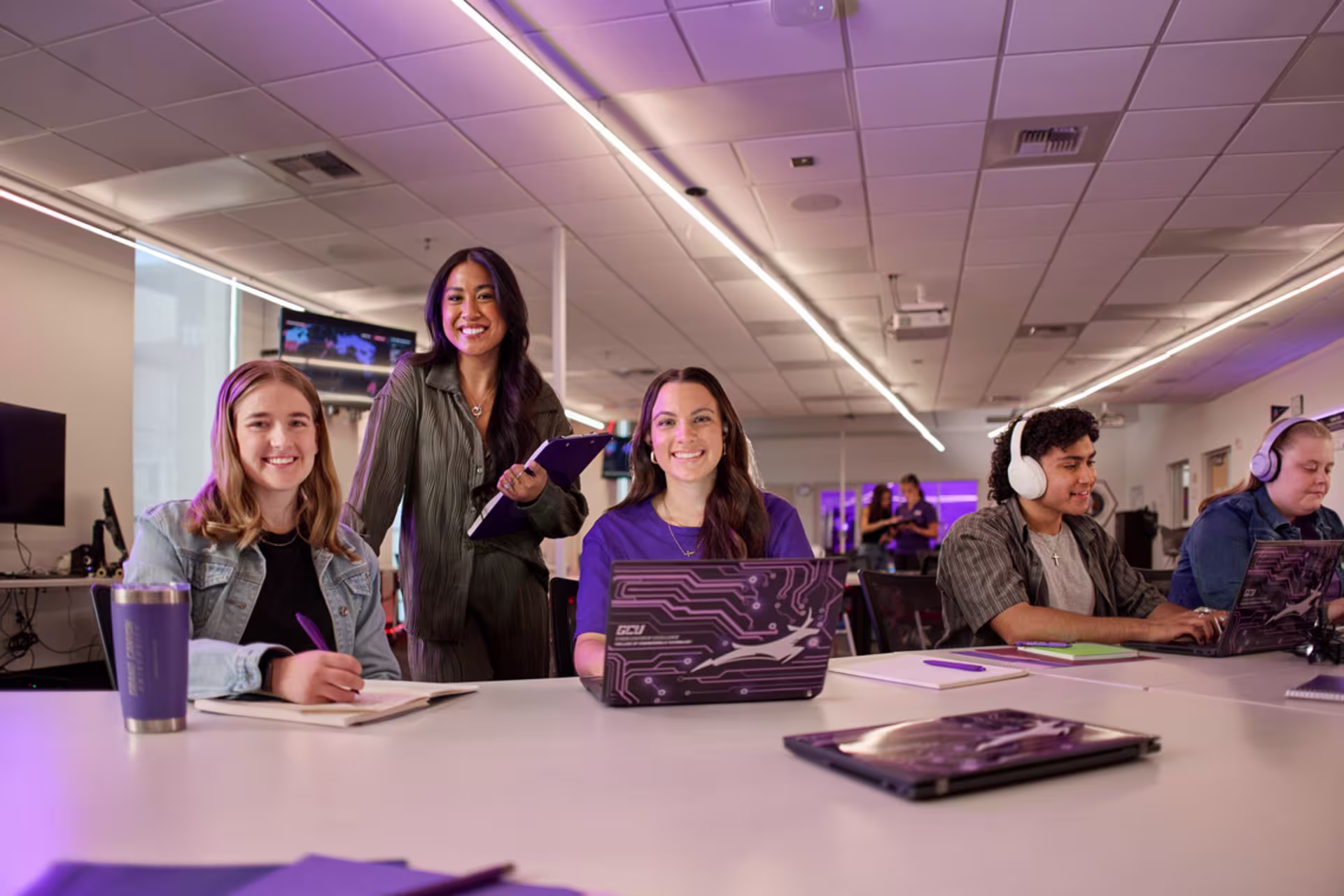 A group of technology degree students works together at a long table with laptops, headphones and notebooks in a computer lab.