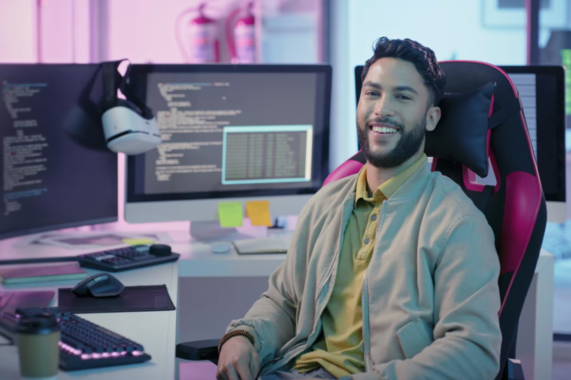 A male web developer smiles at the camera while working at a multi-monitor workstation with code displayed on the screen.