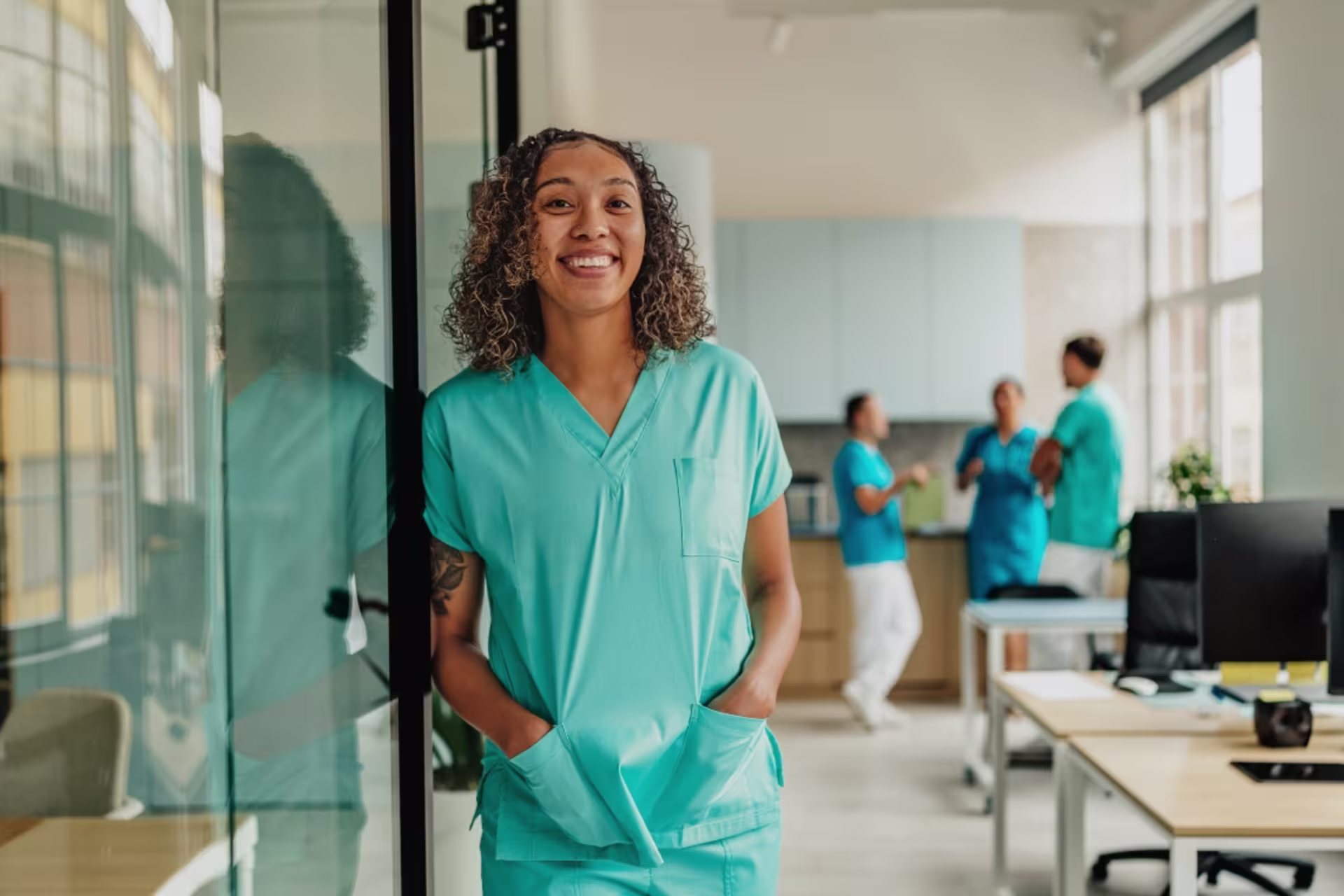 A nurse in teal scrubs smiles while standing in a modern healthcare clinic with medical staff in the background.