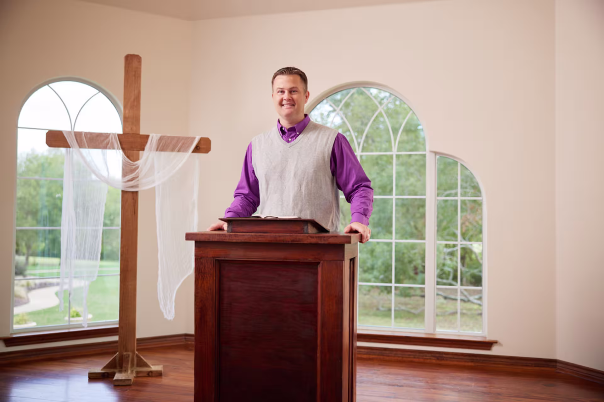 A smiling pastor in a purple shirt and grey sweater vest stands at a pulpit in the front of a church.