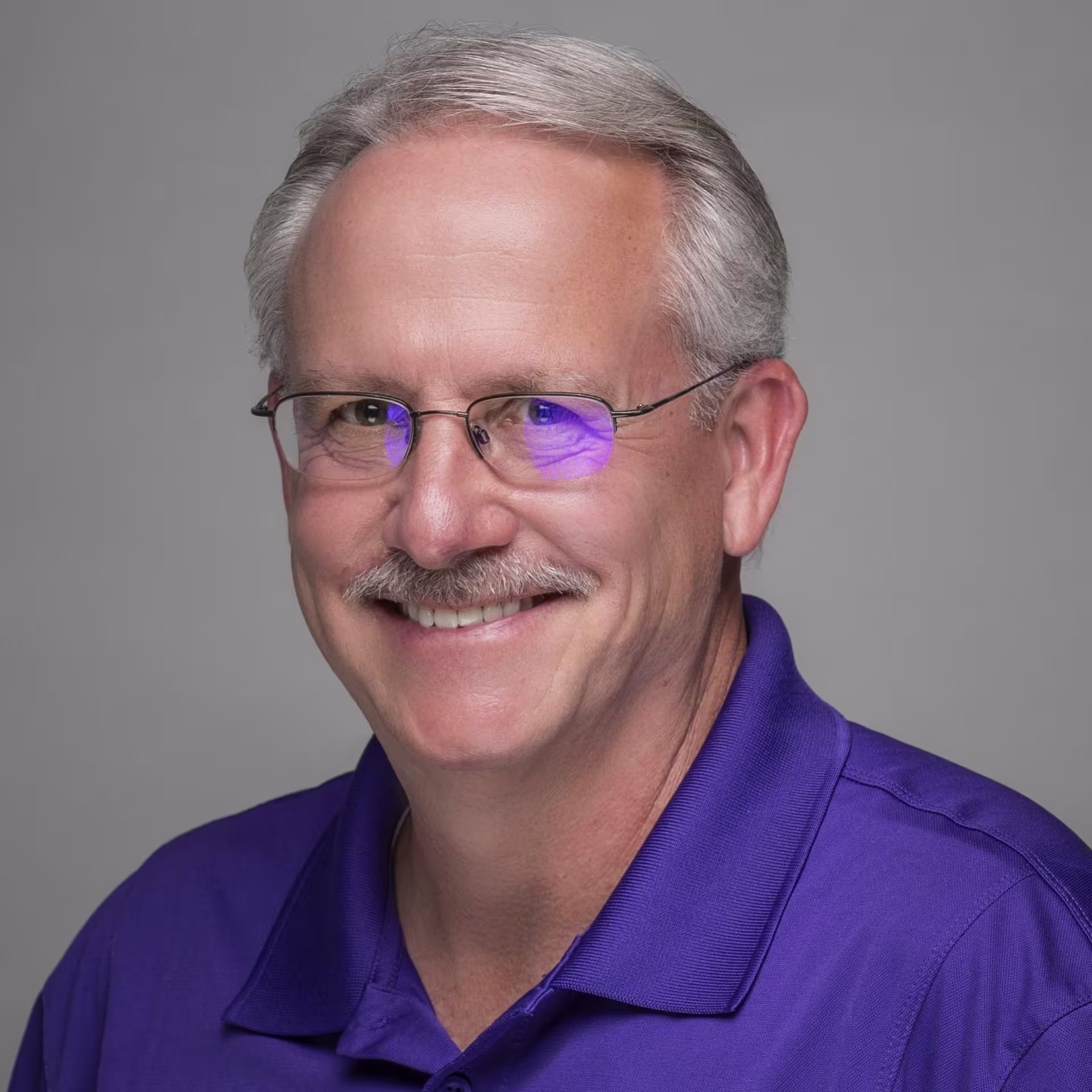 Headshot of GCU faculty member Bill Hughes smiling, wearing glasses and a purple polo shirt against a neutral gray background.