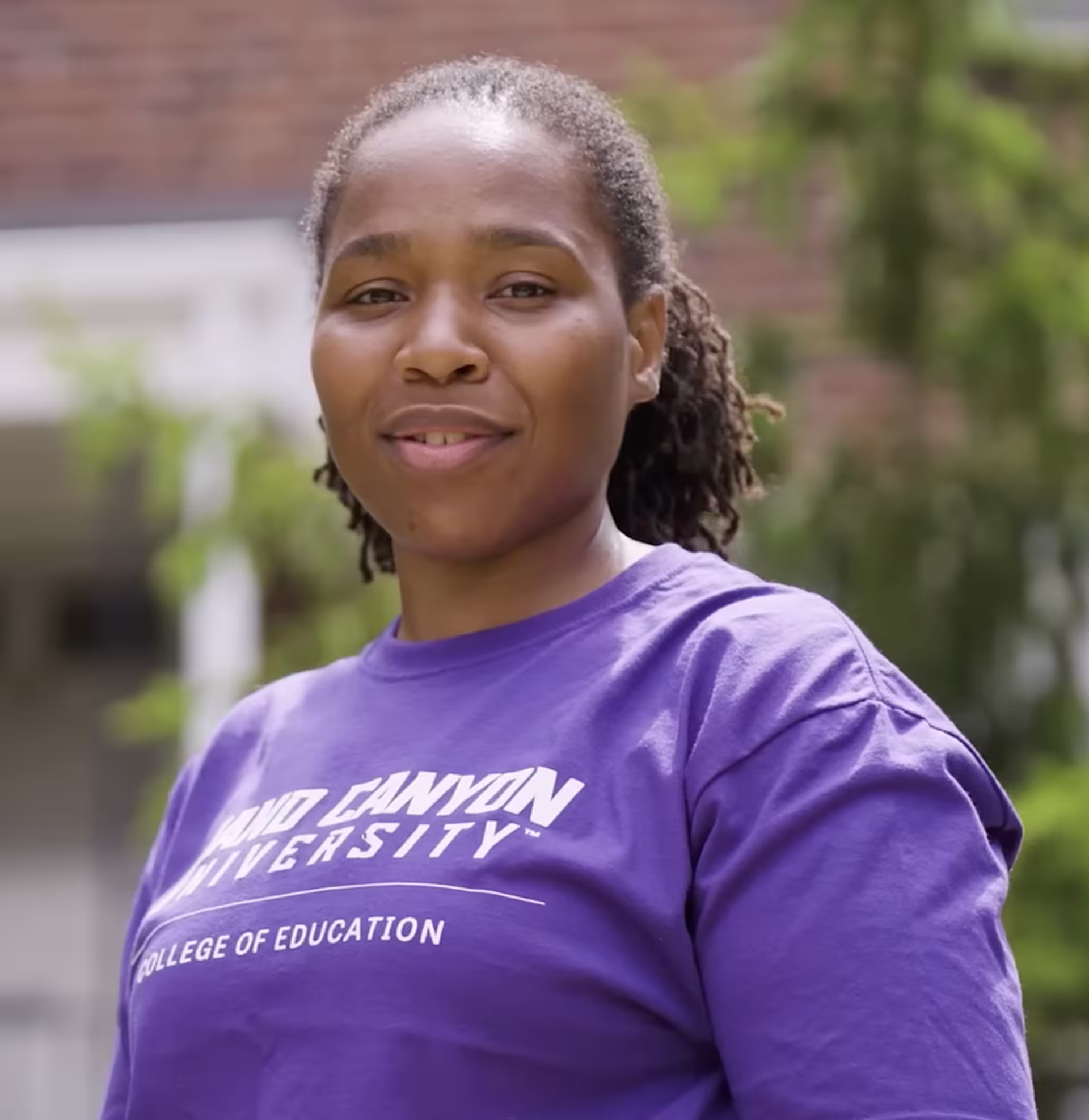 Keisha Ritchie stands outdoors wearing a purple Grand Canyon University College of Education T-shirt, with greenery and campus buildings softly blurred in the background.