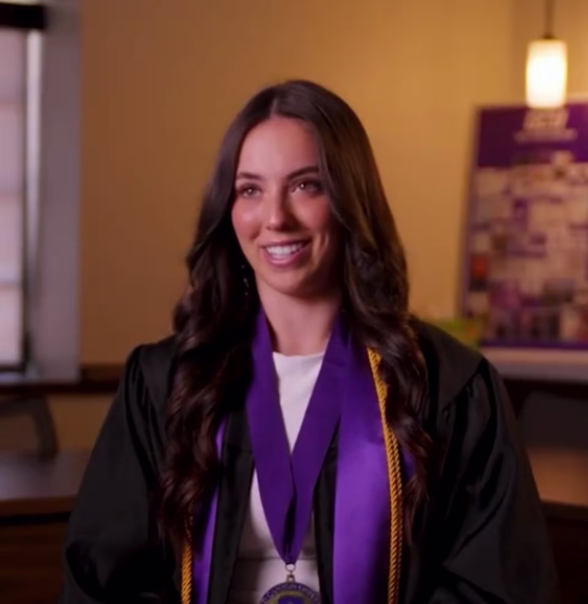 Paige Heilig is seated indoors wearing a black graduation gown with a purple honor stole and medal, positioned against a warmly lit background with university-themed decor.