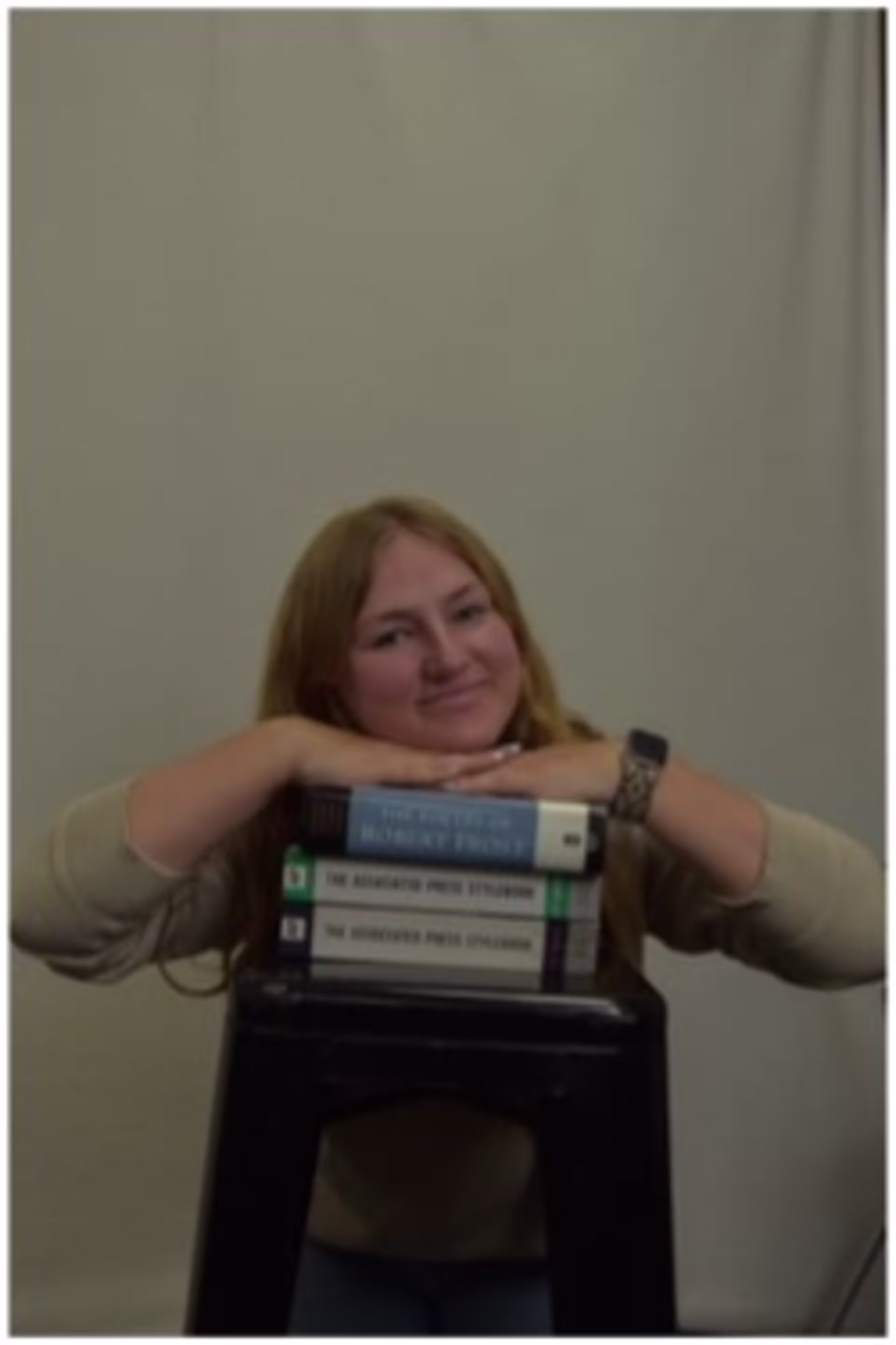 Headshot of Lydia Voxland, professional writing for new media 2026 graduate, seated indoors with hands resting on stacked books.