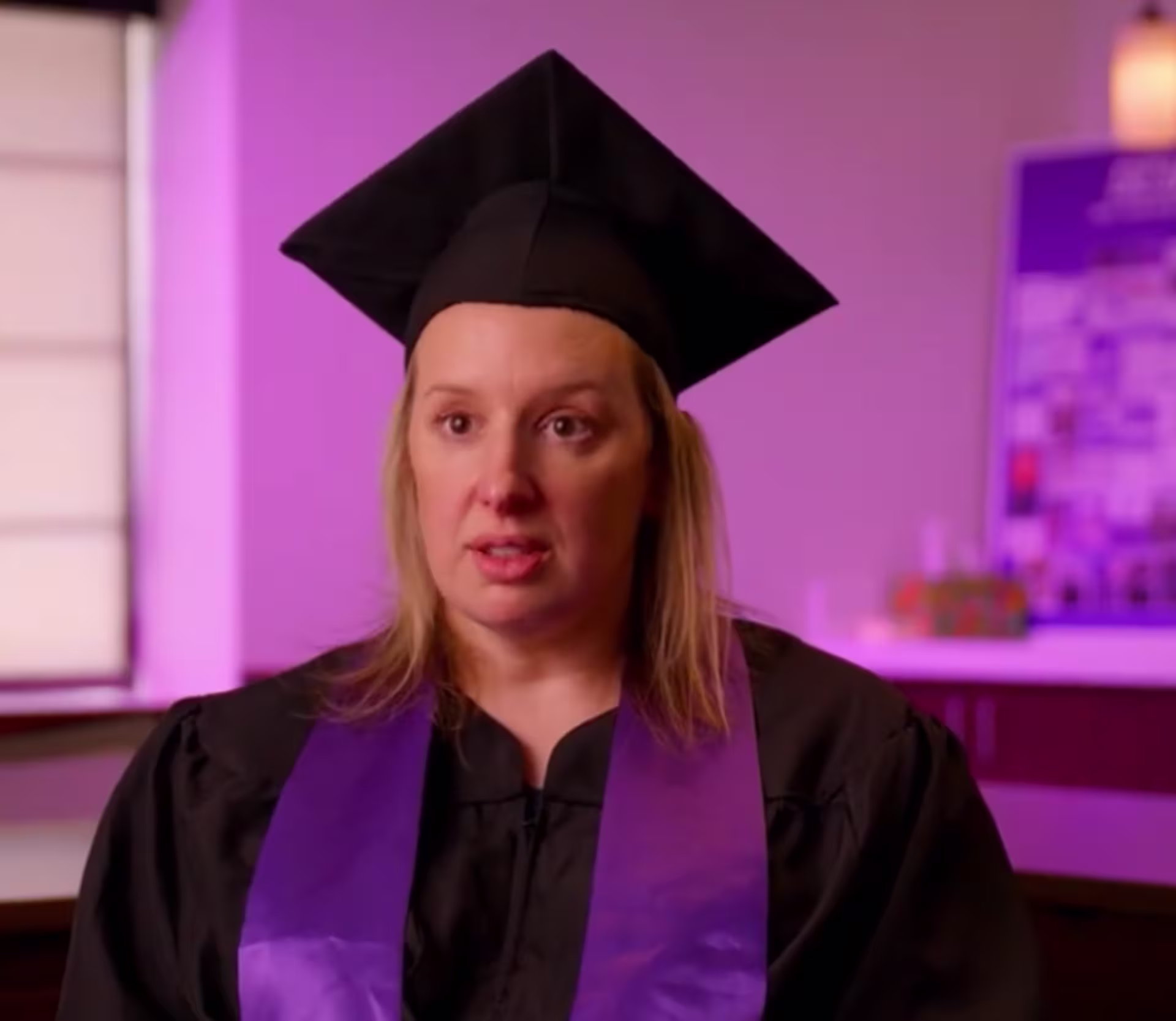 Person wearing a black graduation cap and gown with a purple stole, seated indoors with a softly lit purple background, facing the camera.