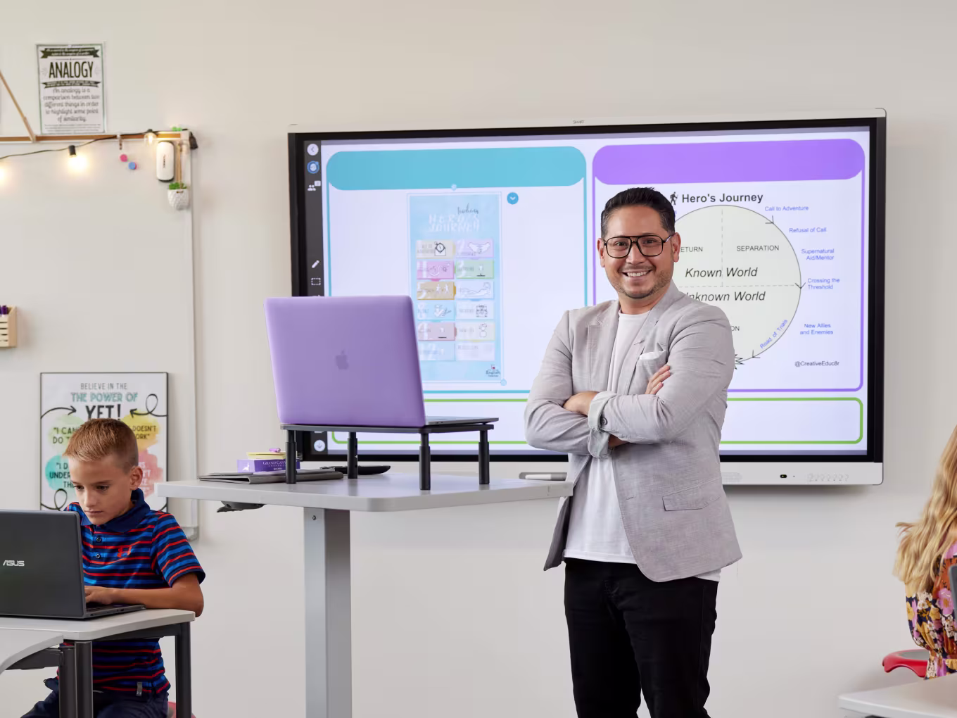 A teacher standing in front of a digital whiteboard explaining the Hero's Journey concept, with students working on laptops in the classroom.