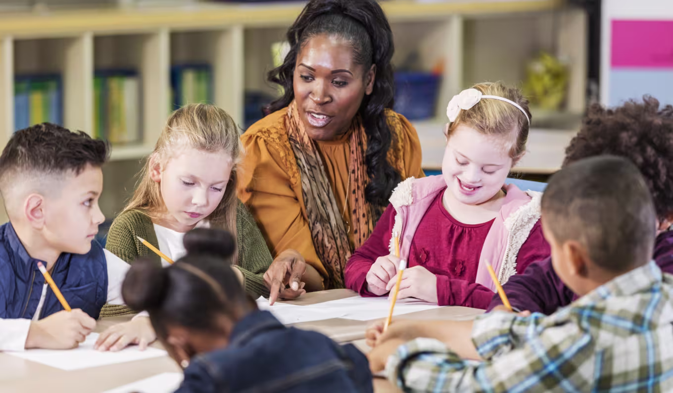 An elementary special education teacher sitting with kids at their classroom table working on their schoolwork.