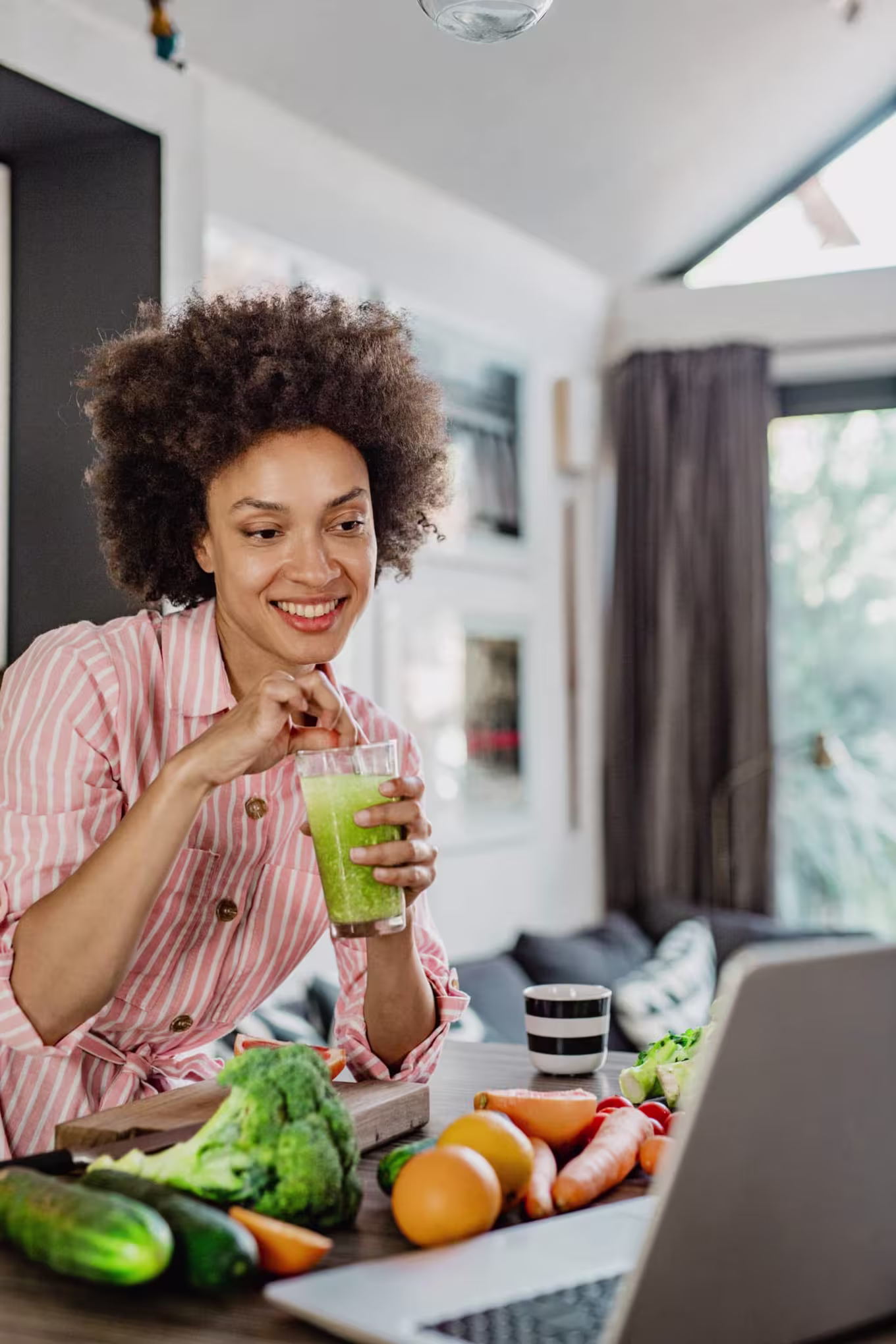 A nutritionist in the kitchen drinking a green juice and studying on laptop.