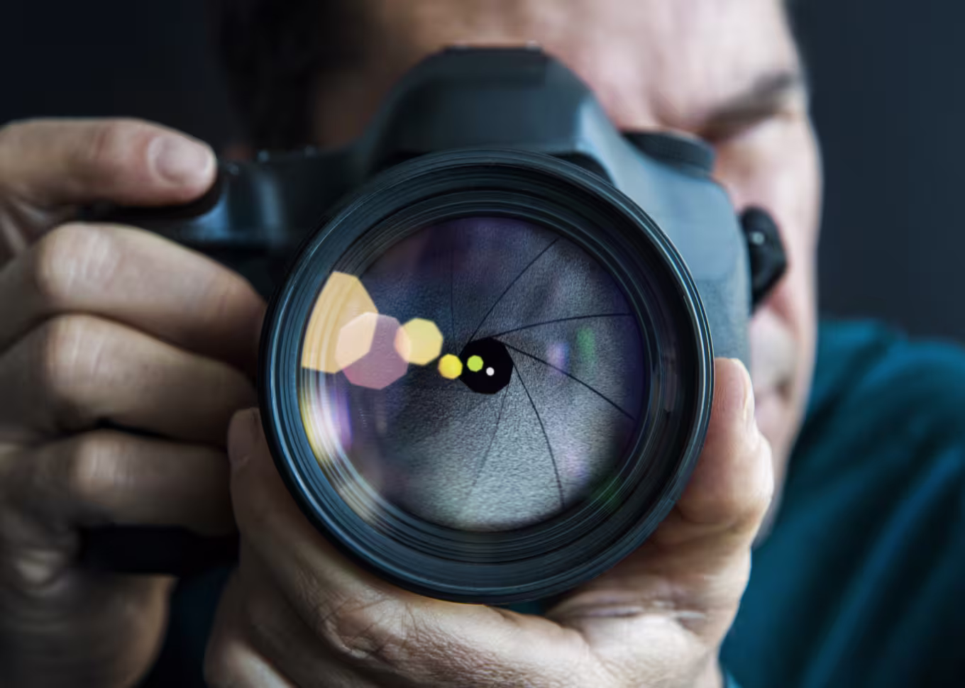 Close-up of a person holding a camera with the lens in sharp focus, showing aperture blades and colorful light reflections.