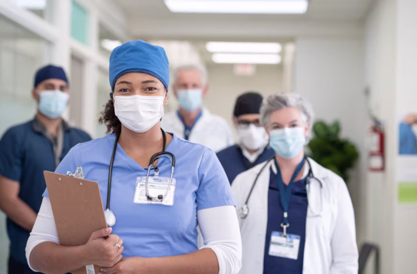 A portrait of a multi ethnic group of healthcare professionals consisting of doctors and nurses at the hospital. A female nurse of African descent is standing in front of the team. Everyone is wearing a face mask and looking at the camera.