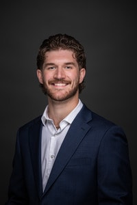 Riley Kollbaum headshot wearing white shirt with navy blue jacket.