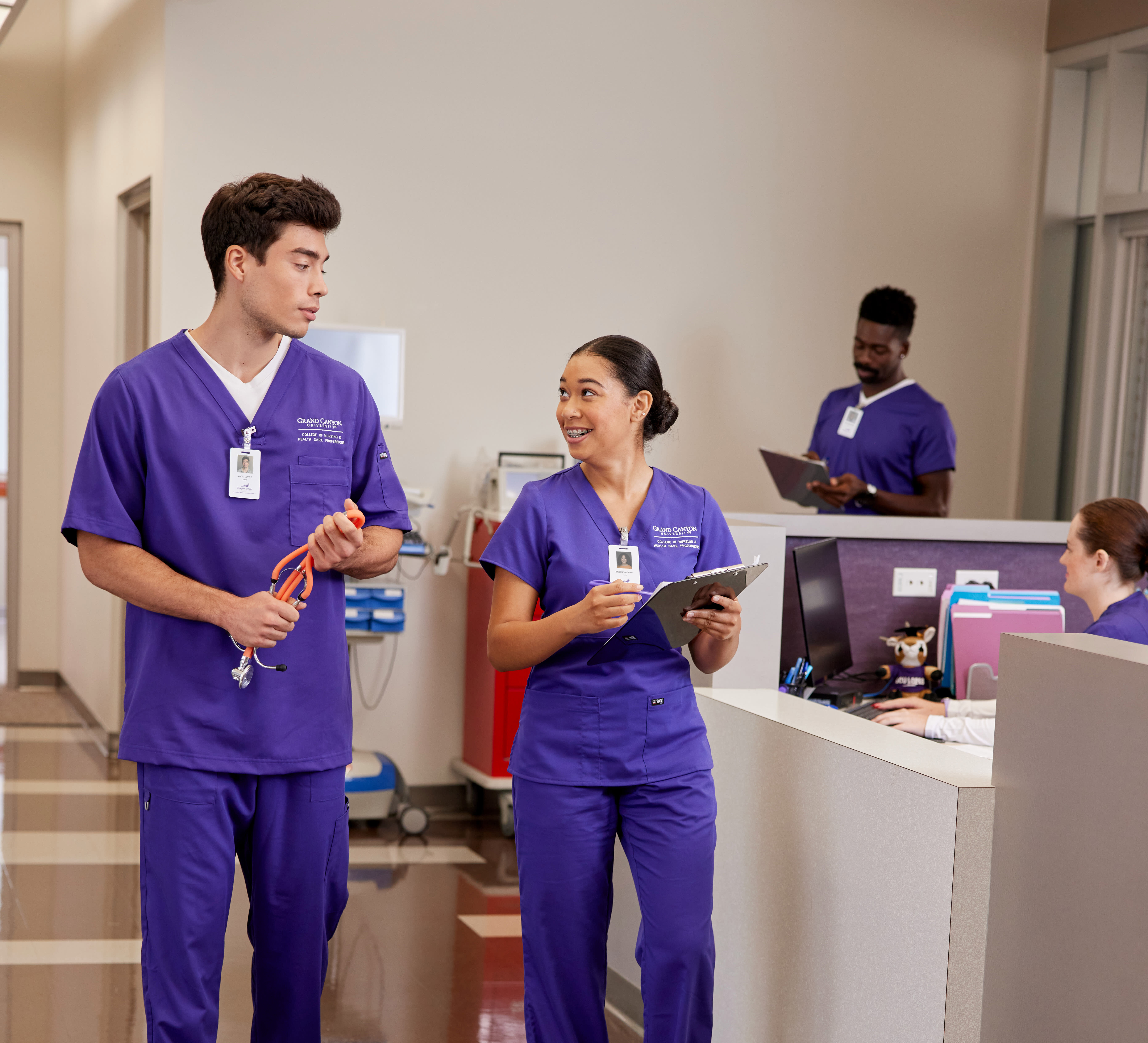 Two Missouri ABSN students in purple scrubs walk past a nurse's station