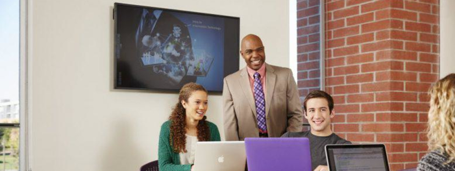 A professor stands behind three students seated and using laptops