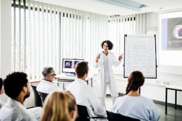 Medical instructor presenting to students in lab coats, discussing strategies for NCLEX exam preparation using visuals.