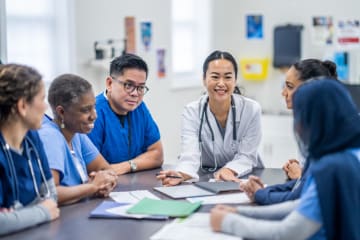 A small group of doctors and nurses gather around a meeting table to discuss patient cases. They are each dressed professionally and have various, files, notes and papers scattered between them as they reference them throughout the meeting.