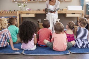 Elementary teacher sits at front of class reading a book to children sitting on ground