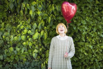 Asian woman with a blonde wig looks up at the heart-shaped balloon she's holding