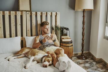Man holds coffee cup looking thoughtfully out the window with dog snuggled up to him on the bed