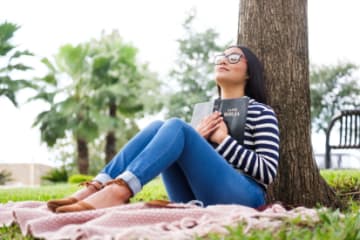 Woman holding an open Bible close to her chest and praying under tree