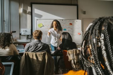 Brunette curly haired teacher stands in front of a white board engaging her students in breaking down a sentence's syntax