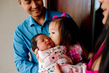 Little Asian girl kissing baby sister on the cheek while man in blue shirt watches