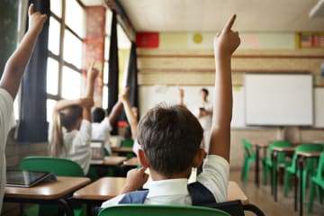 young male student raising hand in classroom alongside other students