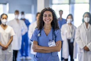 A large group of medical professionals stand in the hallway of a Hospital as they pose together for a portrait. They are each dressed professionally in scrubs and lab coats and some are wearing medical masks to protect them from airborne viruses.