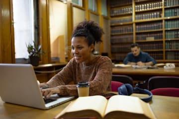 Happy student working in a library with an open book and laptop