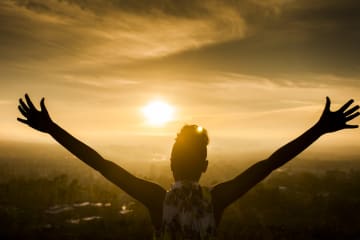 A young African American Woman raises her arms facing the sunset down over the valley and the ocean.