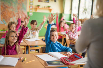 young school children all smiling and raising their hand for the teacher