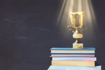 Golden trophy on pile of books, against blackboard, with sun rays over trophy; learning/achievement concept - stock photo