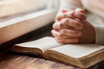 Hands clasped in praying position over open bible next to windowsill