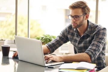 Young male sits at a table working on laptop with coffee and notebooks nearby