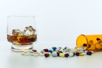 Different pills spill out of bottle next to an alcoholic beverage on ice