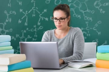 Woman wearing glasses works on laptop with chemistry equations on chalkboard behind her