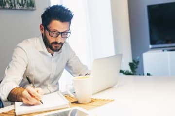 man writing on notebook with laptop in front of him