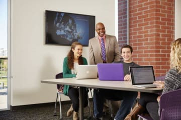 A professor stands behind three students seated and using laptops